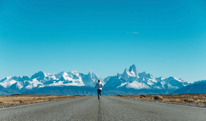 A solo traveler running down a paved road toward snow-capped mountains